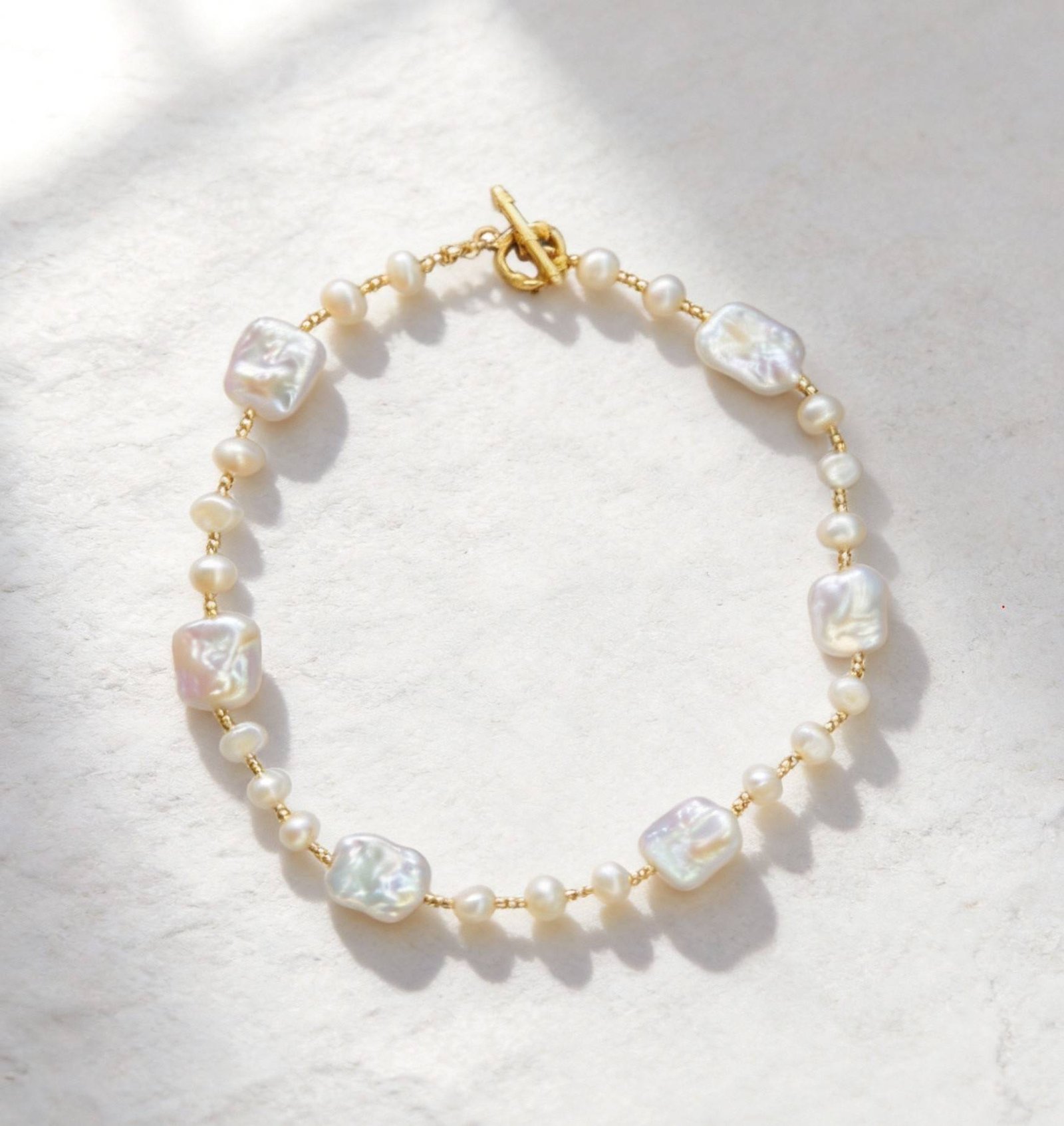 A delicate bracelet featuring baroque pearls and small round pearls on a gold chain with a toggle clasp, on a light stone surface